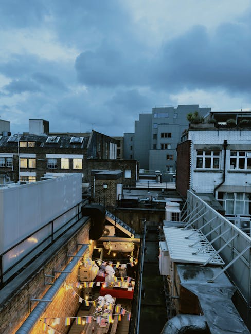 A view from a rooftop outdoor space during dusk shows string lights illuminating a small area with various items, such as cardboard boxes, plastic containers, and loose packaging materials. The scene includes neighboring buildings with diverse architectural styles, including traditional brickwork and modern white façades, with some windows lit from within. Exposed pipes and a metal railing are visible on the rooftop, and the sky above is cloudy with a hint of blue. This urban setting presents elements typical of a house or office relocation project in progress, with [COMPANY_NAME] potentially managing the logistics, including packing and transport of furniture and boxes within the city environment.
