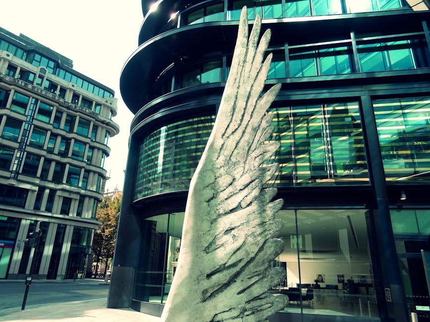 A large, vertical stone sculpture of abstract, wing-like shapes stands on a paved city street in front of a modern glass office building with curved black exterior walls. The sculpture resembles folded or feathered wings and is made of light-colored stone with a textured surface. On the left, there is another contemporary building with a grid-like facade of windows and concrete framing. The area appears well-lit with natural daylight, and the scene is captured during daytime, with a clear sky and some trees visible in the background. This image is associated with Man With a Van Finsbury, a company providing removals and relocation services, and depicts the urban environment where one might perform furniture transport or home relocation tasks involved in a professional moving process.