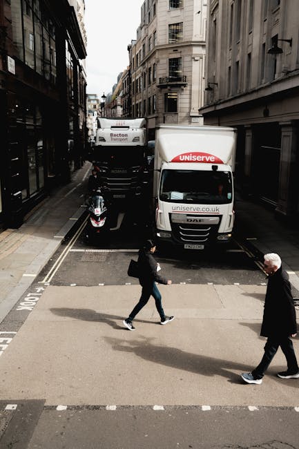 A street scene showing a man with a black backpack and a woman in a dark coat crossing a pedestrian crossing in front of parked delivery trucks and a motorcycle on the street. The trucks are branded w