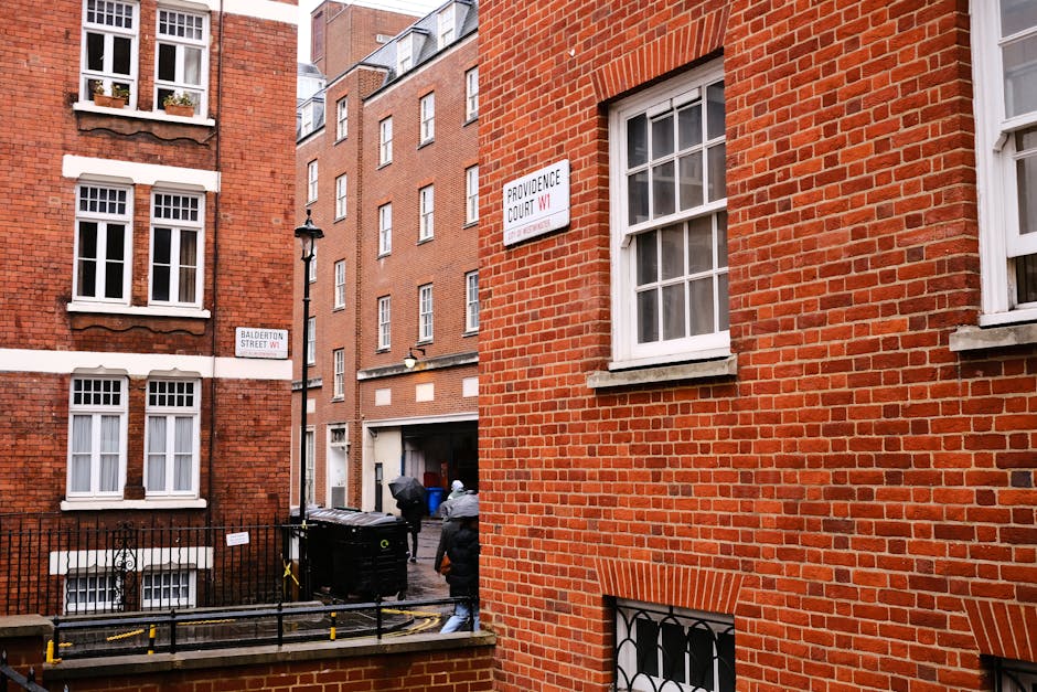 Image depicts a narrow urban residential alleyway with red brick buildings on either side, adorned with white-framed sash windows. A street sign indicates the intersection of Providence Court WI and Balderton Street WI. In the foreground, two movers from Man With a Van Finsbury are loading a large black plastic bin onto a paving area, with one individual seen from behind wearing a grey jacket and white hard hat. The scene includes a black metal fence, a street lamp attached to one of the brick buildings, and a partially visible black van parked nearby. Packing materials such as cardboard boxes and protective blankets are visible around the loading area, suggesting a home relocation process involving furniture transport through a narrow access point. The environment is lit by natural daylight, emphasizing the urban setting and the logistical aspect of the moving service.