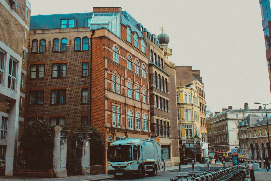 Image depicts a narrow urban residential alleyway with red brick buildings on either side, adorned with white-framed sash windows. A street sign indicates the intersection of Providence Court WI and Balderton Street WI. In the foreground, two movers from Man With a Van Finsbury are loading a large black plastic bin onto a paving area, with one individual seen from behind wearing a grey jacket and white hard hat. The scene includes a black metal fence, a street lamp attached to one of the brick buildings, and a partially visible black van parked nearby. Packing materials such as cardboard boxes and protective blankets are visible around the loading area, suggesting a home relocation process involving furniture transport through a narrow access point. The environment is lit by natural daylight, emphasizing the urban setting and the logistical aspect of the moving service.