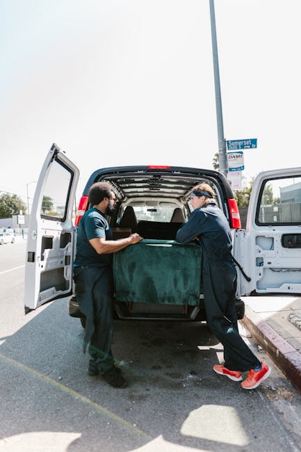 Two movers from Man With a Van Finsbury are engaged in a furniture loading process at a residential street, standing behind an open van door. They are lifting a large, green, padded blanket-covered item, possibly a piece of furniture or appliance, into the vehicle. The van is parked on the roadside near a curb with a purple-painted edge, with its interior empty except for the parcel being loaded. The background includes a street sign indicating the location as Somerset Road and a clear sky above. The scene captures the careful handling and secure transportation involved in home relocation and furniture transport services, with the movers using appropriate packing materials such as blankets to protect the items during loading and transit.