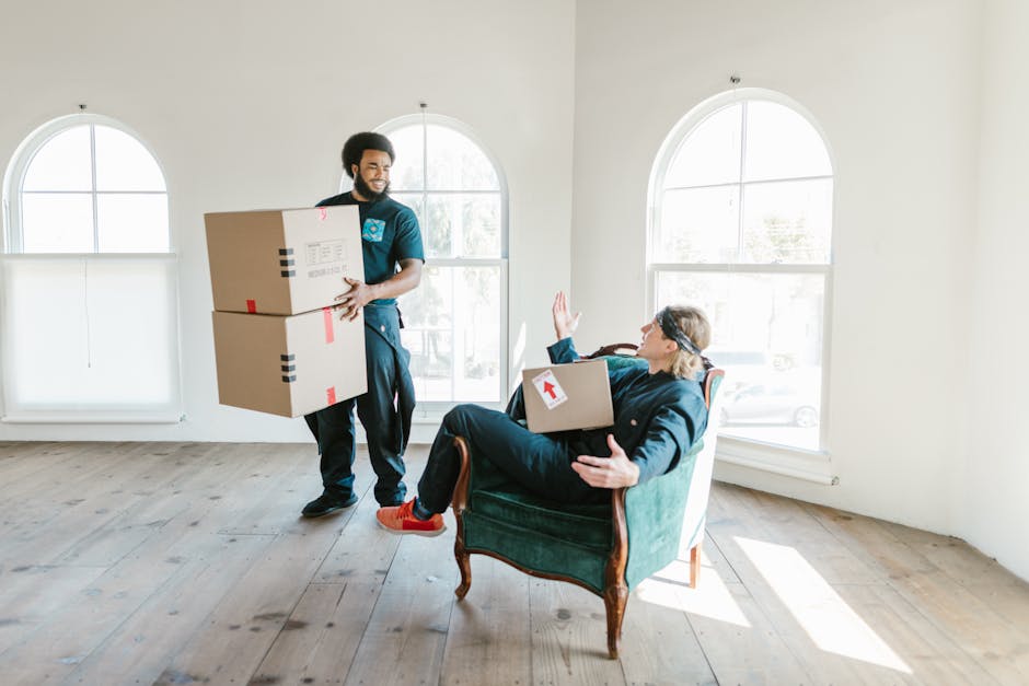A man with dark, curly hair and a beard, dressed in a navy blue uniform with a logo, is carrying two large cardboard boxes taped with red and black packing tape, walking inside a bright, minimally furnished room with light wooden flooring and white walls. Two tall, arched windows in the background let in natural light, illuminating the scene. The woman seated in a vintage-style green armchair, wearing glasses, a dark jacket, and red shoes, is holding a cardboard box with a red upward arrow sticker, and raising her hand in a gesture of greeting or acknowledgment. The room appears to be in the process of packing or unpacking, with some empty space and no visible furniture or other items, suggesting that [COMPANY_NAME] is assisting with a home relocation or furniture transport. The environment is clean and well-lit, emphasizing the logistic and packing steps involved in a house removal or moving service.