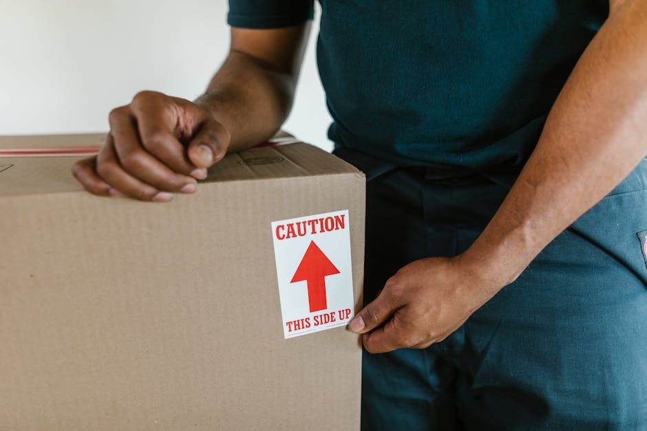 A person wearing a dark green shirt and blue work pants is holding a large cardboard box, which has a white label with red text and an arrow indicating the correct orientation, reading 'Caution: This Side Up'. The individual is gripping the box at its sides, with one hand positioned towards the top and the other towards the bottom. The box is inside a property, possibly on a floor or table, ready for a home relocation or furniture transport process. In the background, a plain, light-colored wall is visible, and the lighting is bright and even, highlighting the focus on careful packing and adherence to proper handling procedures during house removals. This image illustrates the importance of correct box orientation during packing to avoid damage or delays in moving services, as emphasized by Man With a Van Finsbury, specialists in removals and moving logistics.