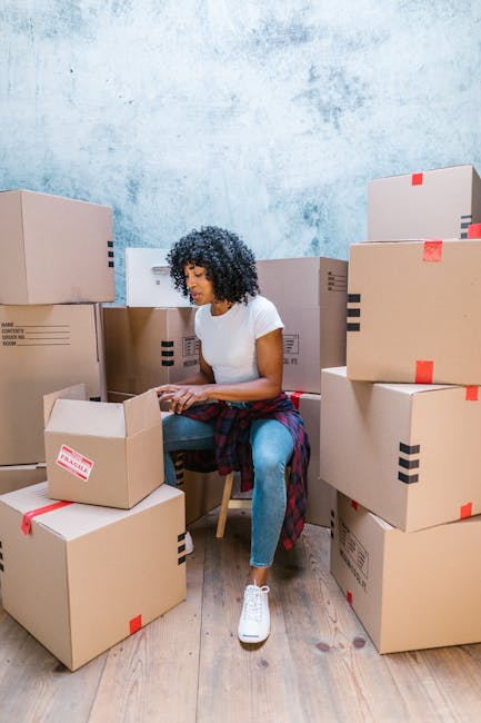 A woman with curly dark hair, wearing a white t-shirt, blue jeans, and white sneakers, is seated on a wooden chair amidst numerous packed cardboard boxes with black and red packing tape. The boxes are stacked and arranged around her, some on the floor and some on top of each other, indicating an active packing process. The setting appears to be an indoor space with a textured blue wall in the background and wooden flooring. She is engaged in packing or organizing items, highlighting the packing and moving stage of a house relocation. This scene is associated with home moving services offered by Man With a Van Finsbury, specialising in removals and same-day or urgent moves, illustrating the preparatory activities involved in a furniture transport or home relocation.