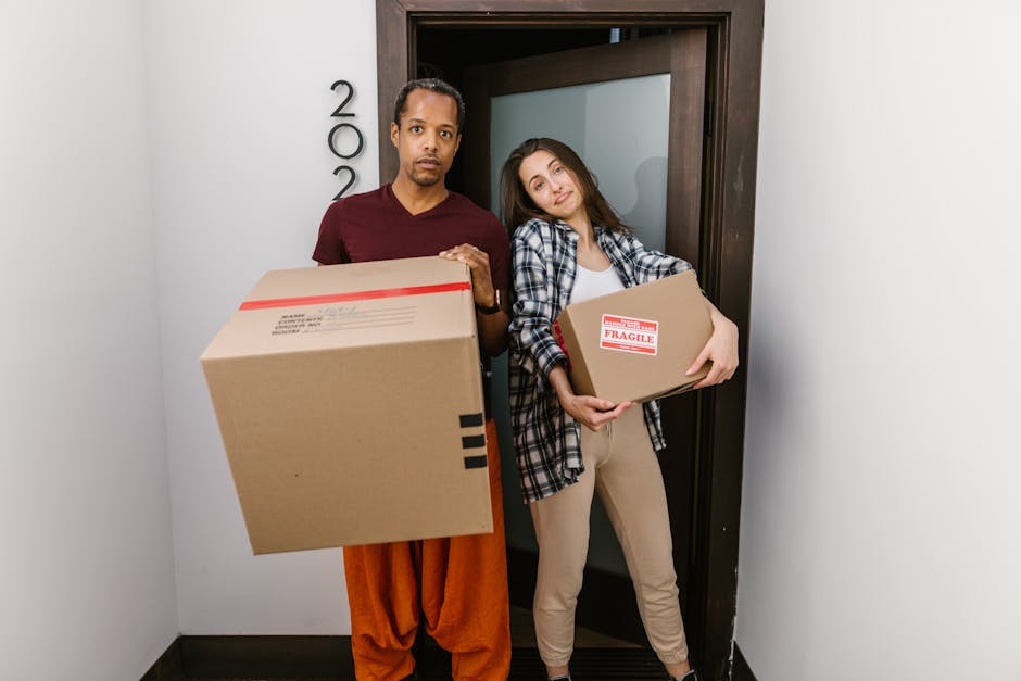A man with a serious expression and a woman with a slight smile stand inside a building near a doorway, each holding a cardboard box. The man, dressed in a maroon T-shirt and orange trousers, is carrying a large box with black tape along the edges, while the woman, wearing a plaid shirt over a white top and beige pants, holds a smaller box with a red 'Fragile' sticker. The boxes appear to be packed for home relocation, part of a furniture transport or packing and moving process. The interior features white walls, a dark door frame, and a partially open frosted glass door. The scene is well-lit, emphasizing the moving activity, which is related to house removals and urgent move options as offered by Man With a Van Finsbury, located in Finsbury and visible in the context of same-day removals services.