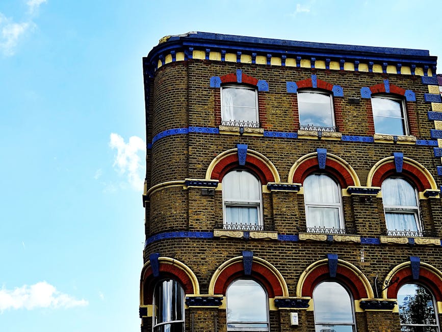 Close-up view of a historic brick building with ornate architectural details and arched windows, surrounded by a clear blue sky with a few white clouds. The building features decorative blue and red b
