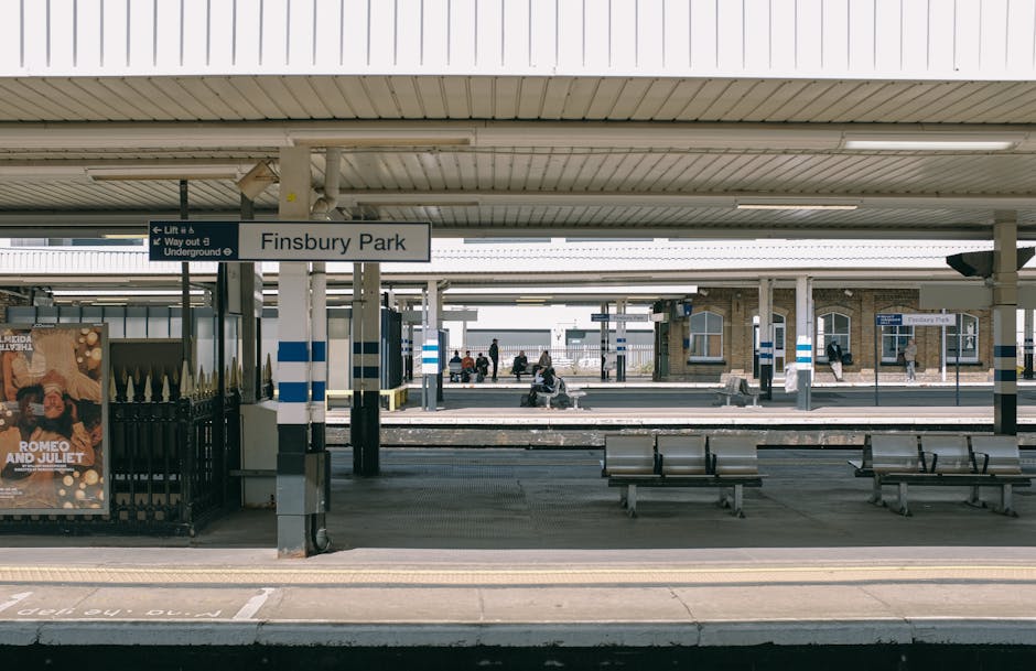 The image shows Finsbury Park train station platform with a white sign displaying the station name in black text suspended under a beige, corrugated metal roof. Several passengers sit on metal benches along the platform, some appearing to wait for a train. On the left side, there is a poster advertising a theatrical production of 'Romeo and Juliet' mounted on a black metal fence. The platform edge features tactile paving, and the surrounding environment includes additional platforms and station infrastructure in the background. The scene is well-lit with natural daylight, and the station appears prepared for home relocation and furniture transport activities, with the station's facilities facilitating loading and unloading luggage or moving boxes, supported by Man With a Van Finsbury’s logistics. This setting naturally ties into the process of packing, loading, and transport involved in house removal services.
