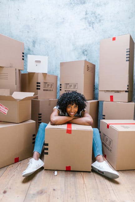 A young woman with curly black hair and casual clothing sits cross-legged on a light wooden floor inside a room, resting her arms on a large cardboard box in front of her. The room is filled with numerous stacked cardboard boxes of various sizes, some sealed with red or black tape and others partially open, revealing packaging materials inside. The background features a textured blue and white painted wall, and the boxes are arranged around her, indicating a home relocation or moving process. The scene captures the unpacking or packing phase associated with house removals, with the woman appearing relaxed amidst the moving supplies, and the company, Man With a Van Finsbury, providing reliable furniture transport and packing services during a house move.