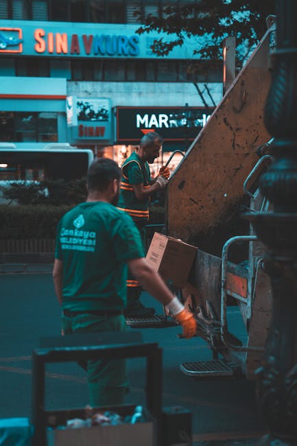 Two workers from Man With a Van Finsbury are loading cardboard boxes and packing materials into the back of a large, open industrial waste collection vehicle, which is parked on a paved area outside a building during twilight. One worker, positioned nearer to the camera, is wearing a teal uniform with the company’s logo and appears to be preparing the contents for transport, while the other, further in the background, is wearing a striped shirt and is actively placing cardboard boxes into the vehicle’s hatch. The vehicle is equipped with a hydraulic lifting mechanism, and there are several stacked cardboard boxes and plastic-wrapped items nearby, suggesting a house removal or moving process involving packing and transporting household belongings. The surrounding environment features signage on nearby buildings, indicating an urban commercial area, and the lighting conditions indicate late afternoon or early evening. This scene illustrates a home relocation or furniture transport operation, possibly related to managing bulky waste disposal during a house move, consistent with regulations for removals in Islington.