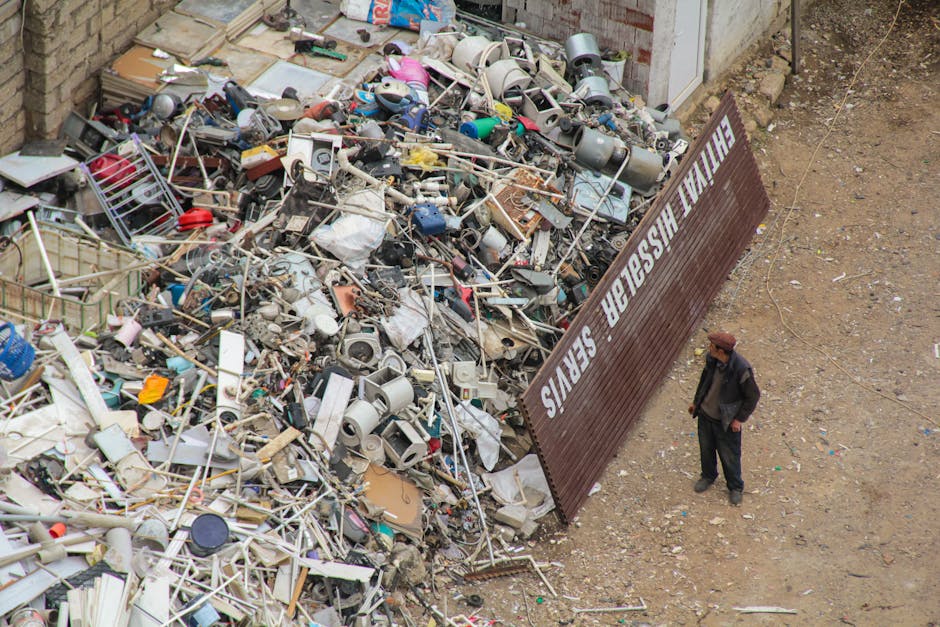 A large pile of assorted household waste and bulky items, including plastic containers, buckets, metal and plastic pipes, broken furniture, and discarded electronic appliances, is situated outdoors on a gravel surface. The waste is situated behind a brown metal fence with the words 'County Waste Services' painted in white, which is partially open. A man wearing dark clothing and a brown hat stands nearby on the right side of the image, observing the waste. The scene is illuminated by natural daylight, with a brick wall visible at the back of the waste pile. The setting appears to be an area designated for waste disposal or recycling, with the waste materials indicating a recent clearing or disposal process involved in home relocation or decluttering activities, supported by the presence of a large quantity of mixed waste materials typical of removals or disposal jobs managed by Man With a Van Finsbury.