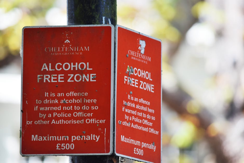 Two red Cheltenham Borough Council 'Alcohol Free Zone' signs are attached to a metal pole on a pavement, with one sign overlapping the other. The signs display white text indicating the zone is designated to prevent alcohol consumption without police authorization, and state the maximum penalty of £500. The signs are weathered, with some scratches and chipped areas, and are positioned outdoors with a blurred background of trees and natural light. The surrounding environment suggests the signs are in a residential or urban area that is subject to local regulations on public alcohol consumption. The scene captures the context of urban signage related to local council rules, relevant to house removals or relocations in the UK.