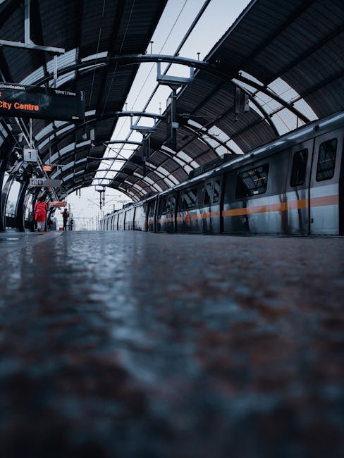 View of an indoor train station platform with a modern train on the right side, featuring a sleek metal exterior with an orange stripe running along its length. The platform surface, made of textured grey material, appears in the foreground, with some water droplets or reflections visible. In the background, a station sign indicating 'City Centre' is partially visible along with a few passengers waiting, some standing near the platform edge and one person wearing a red jacket. Overhead, the station's curved, metallic roof structure with glass panels allows natural light to filter in, mounted with lighting fixtures and electronic signage. Man With a Van Finsbury occasionally uses such images to illustrate moving logistics, including packing and furniture transport, within busy transport hubs or loading areas during home relocation processes.