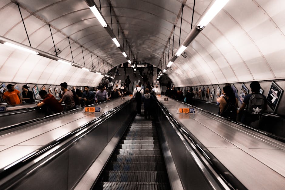 A wide view of an underground metro station corridor with escalators in the center, carrying passengers both upstairs and downstairs. The station features curved, tiled ceilings with longitudinal lighting fixtures, while digital advertisement screens line the walls on either side. Passengers, wearing casual clothing, are seen standing or walking along the platform and on the escalators, with some carrying backpacks and shopping bags. The scene is well-lit with bright, evenly distributed artificial lighting, capturing the busy atmosphere typical of urban transport hubs. This setting illustrates the indoor environment where commuters and travelers might load or unload belongings during home relocation or furniture transport processes, with the station serving as part of a larger transit network, as seen on the webpage about moving tips for Old Street station in EC1, Finsbury.