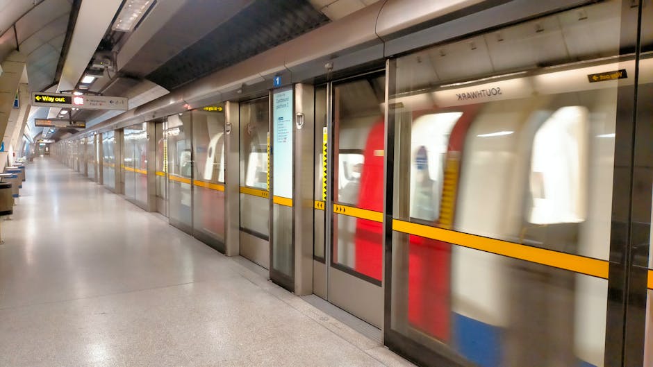 View of an indoor train station platform with a modern train on the right side, featuring a sleek metal exterior with an orange stripe running along its length. The platform surface, made of textured grey material, appears in the foreground, with some water droplets or reflections visible. In the background, a station sign indicating 'City Centre' is partially visible along with a few passengers waiting, some standing near the platform edge and one person wearing a red jacket. Overhead, the station's curved, metallic roof structure with glass panels allows natural light to filter in, mounted with lighting fixtures and electronic signage. Man With a Van Finsbury occasionally uses such images to illustrate moving logistics, including packing and furniture transport, within busy transport hubs or loading areas during home relocation processes.